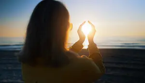 Mature woman accessing spiritual benefits of the beach by framing the sun with her hands.