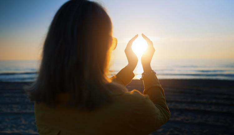 Mature woman accessing spiritual benefits of the beach by framing the sun with her hands.