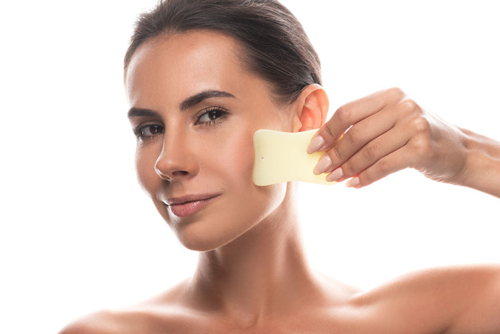 A woman uses gentle scraping with a smooth jade tool for Gua Sha treatment on her face.